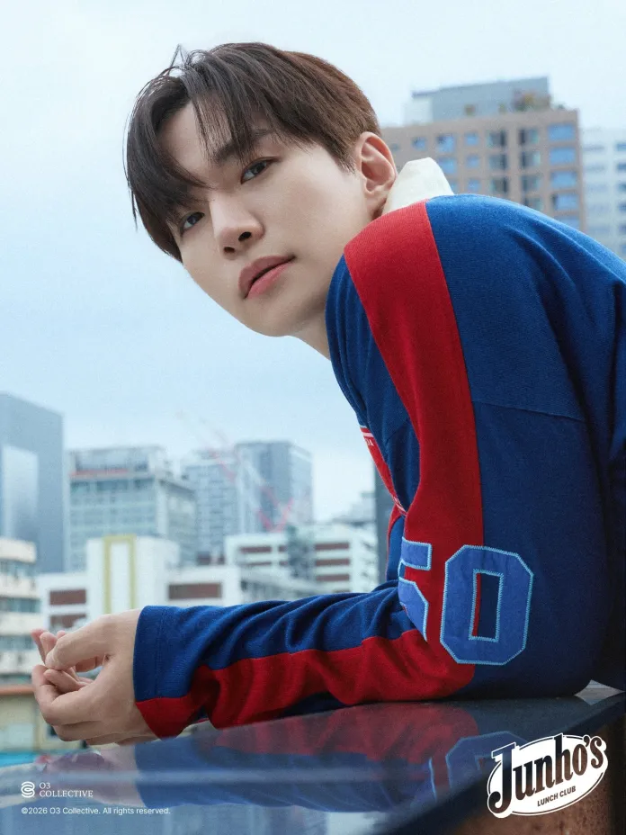 Lee Junho with brown hair wearing a blue and red sweatshirt leans against a glass railing, gazing thoughtfully towards the camera, with a modern cityscape in the background.