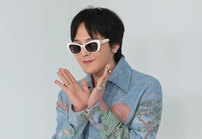 G-Dragon wearing white sunglasses and colorful clothing, striking a playful pose with their hands together. The background is light-colored.