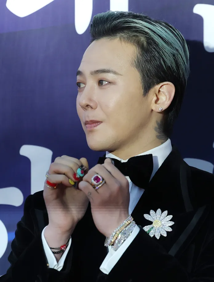 G-Dragon with styled hair, wearing a tuxedo with a bow tie and a decorative flower pin. He is showing colorful nail art on his hands adorned with multiple rings.