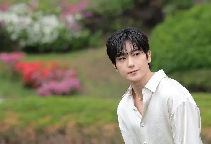 Hyunjae with dark hair wearing a light-colored button-up shirt, standing in a garden with colorful flowers in the background.