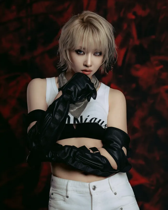 Minzy with short blonde hair wearing a sleeveless white top and black leather gloves, looking directly at the camera with a serious expression. The background features a dark, abstract pattern.