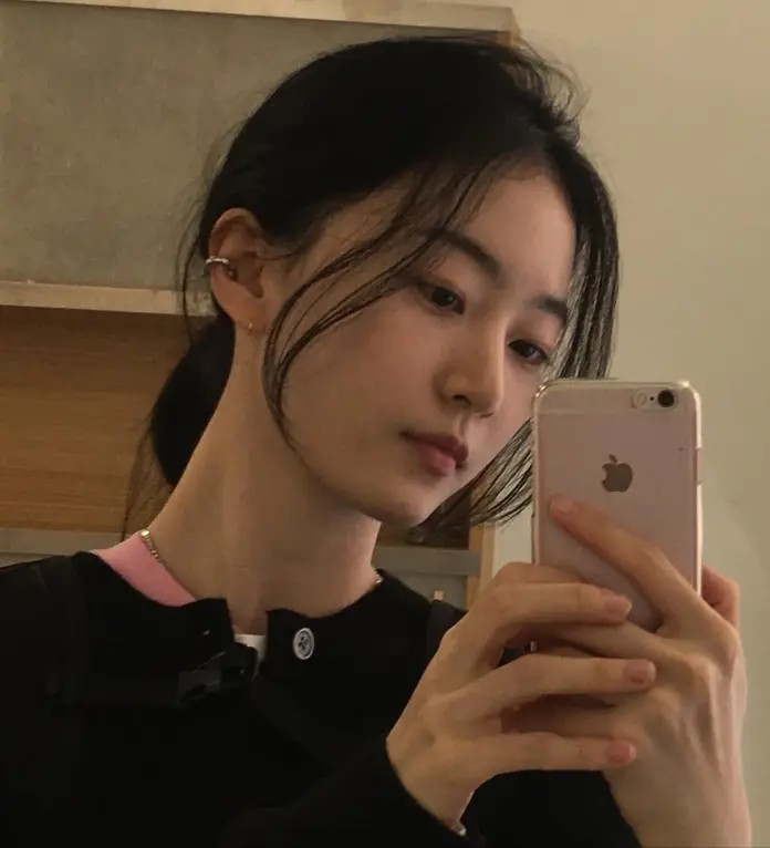 A young woman with long dark hair holds a smartphone, taking a selfie in a modern setting. She appears thoughtful, with a slight smile and a casual black outfit.