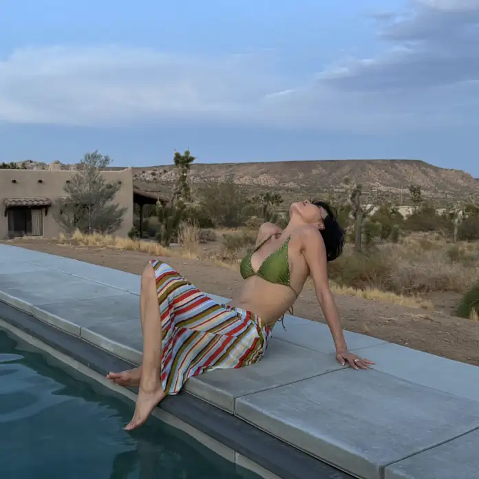 Hwasa in a green bikini and a colorful striped skirt sitting on the edge of a pool, leaning back and enjoying the view of a desert landscape with mountains in the background.