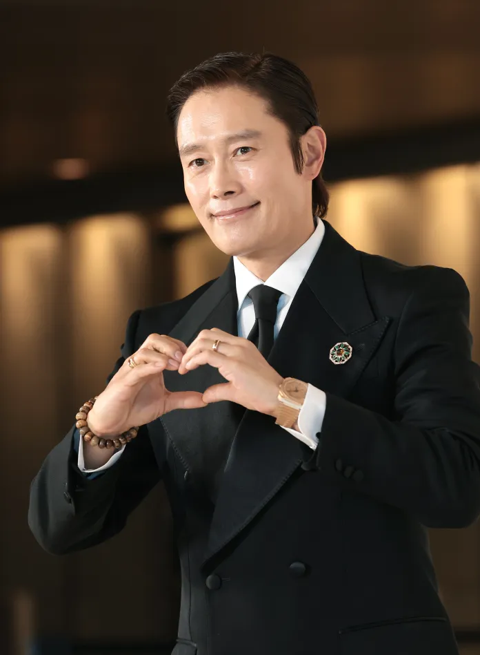 A man in a black tuxedo making a heart shape with his hands, smiling at the camera.