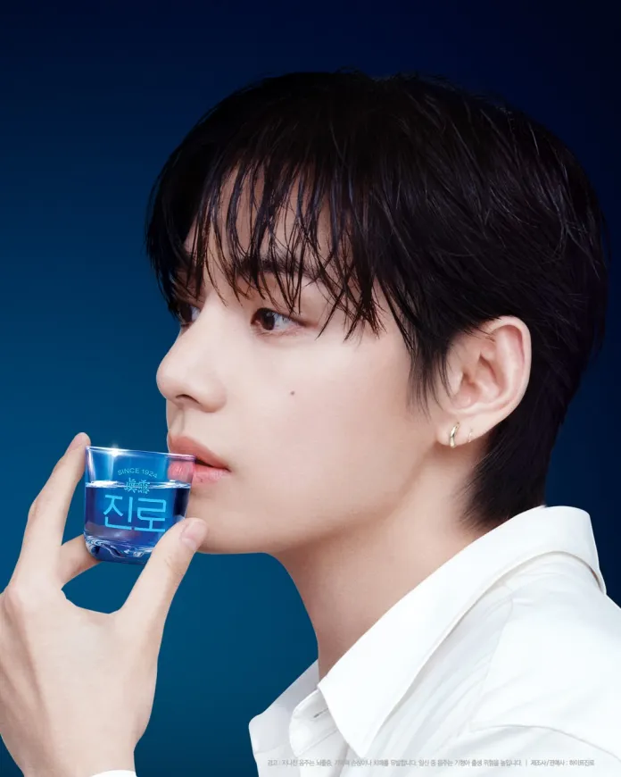 V with short, wet hair holding a small glass of blue liquid, set against a dark blue background.