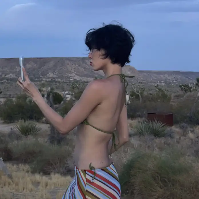 Hwasa in a bikini stands in a desert landscape, holding a phone and looking at it, with mountains in the background.