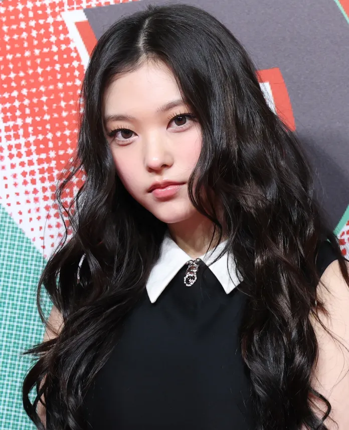 A young woman with long, wavy black hair wearing a black dress with a white collar, posing with a serious expression against a colorful background.
