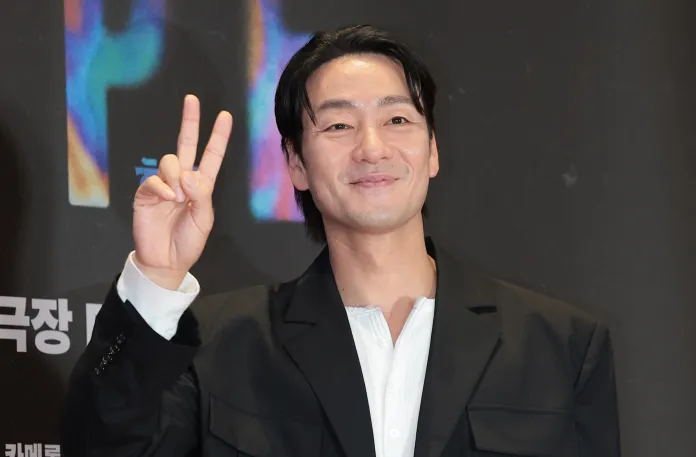 A man wearing a black blazer and a white shirt smiles and holds up a peace sign at a promotional event.