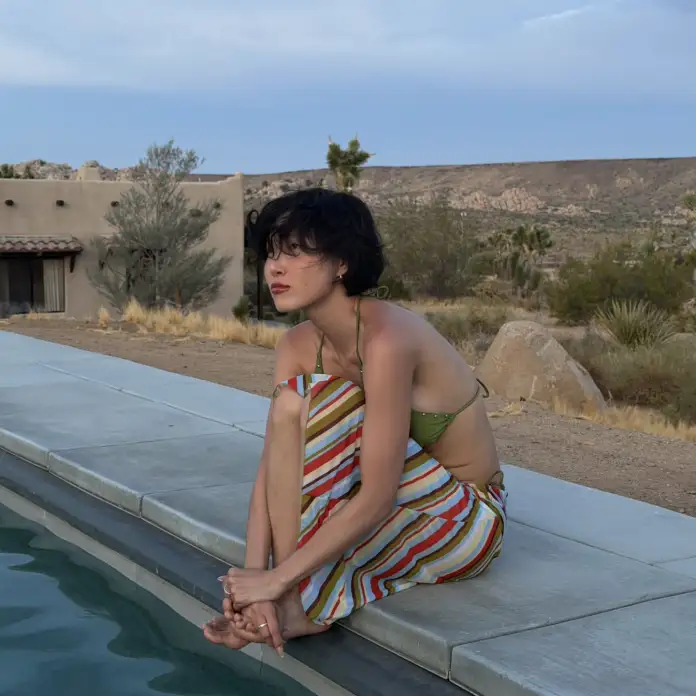 Hwasa sitting by a swimming pool with a contemplative expression, wearing a green bikini top and a multicolored striped skirt, surrounded by a desert landscape.