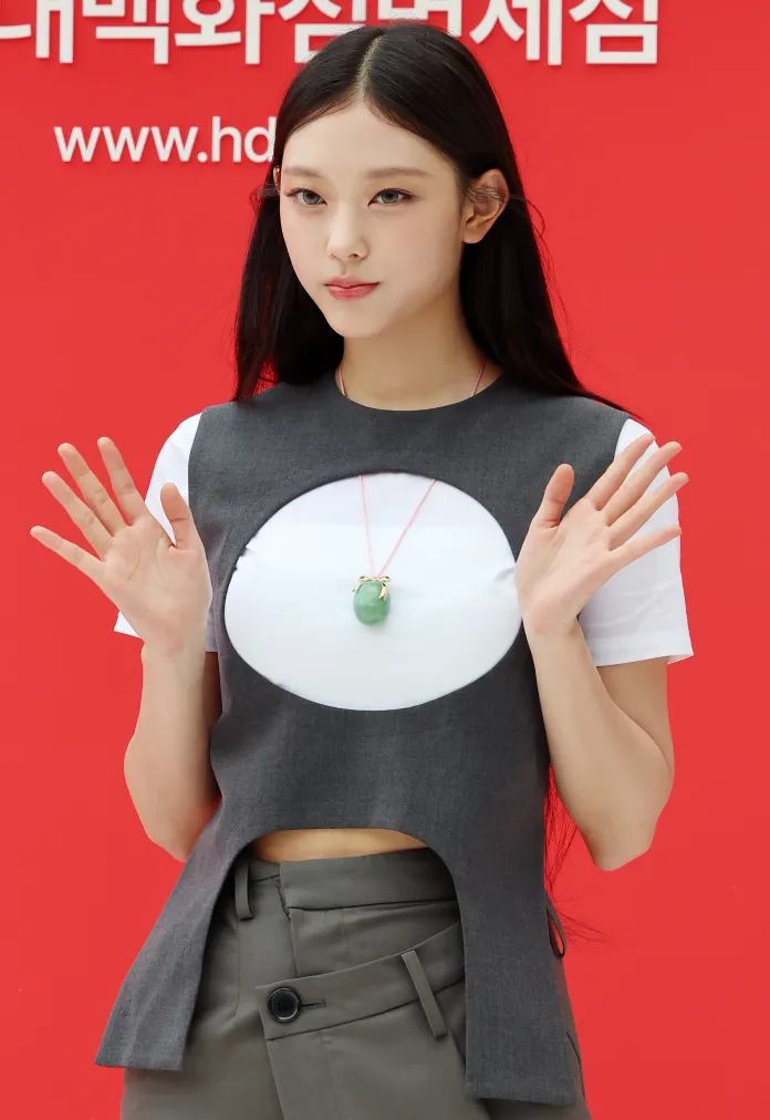 A young woman with long black hair is posing with her hands raised, wearing a unique gray sleeveless top over a white shirt and green pants, standing against a red background.