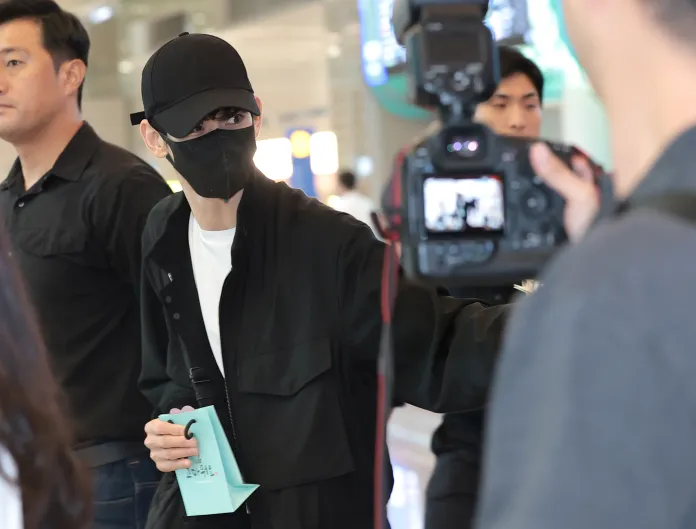 Kim Soo-hyun wearing a black cap and face mask holding a light blue bag, surrounded by individuals in a public space, with a camera pointed at them.