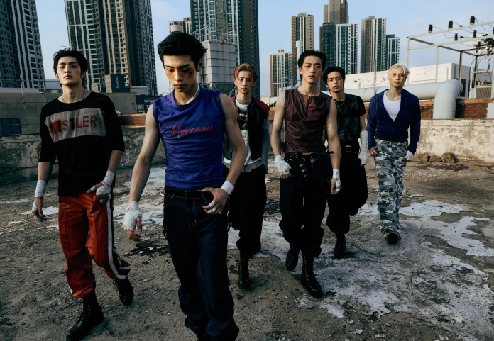 A group of six male models walking together on a rooftop, wearing stylish urban outfits. The skyline of tall buildings is visible in the background.