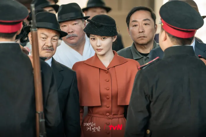 Kim Yoo-jung in a stylish orange coat and black hat surrounded by several men in dark suits and hats, with a tense atmosphere signaling a confrontation.