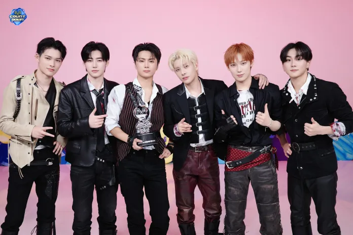 A group of six young men posing together in front of a pink background, dressed in stylish, coordinated outfits. One member is holding an award trophy.