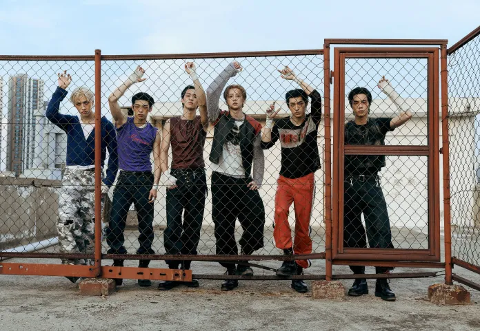 A group of seven young men posing against a chain-link fence, showcasing a diverse range of fashion styles and expressions.