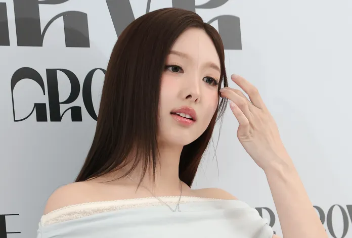 Nayeon with long, straight hair is touching their face while standing against a backdrop with the word 'GROW' in bold letters. They appear to be in a poised, thoughtful stance.