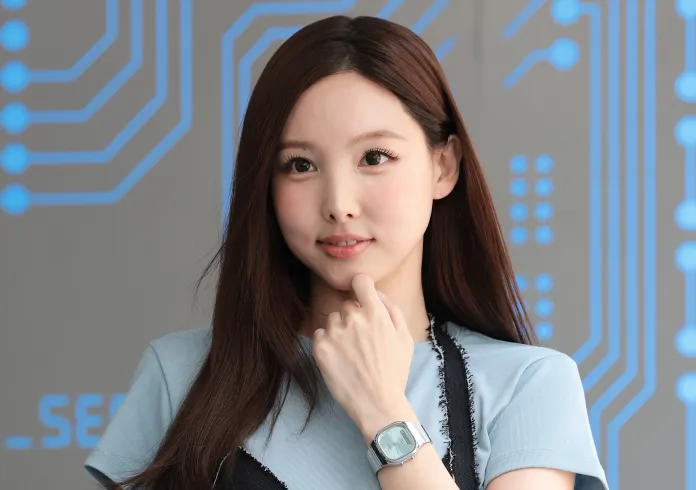 Nayeon with long brown hair wears a light blue shirt and a stylish watch, posing thoughtfully in front of a circuit-themed backdrop.
