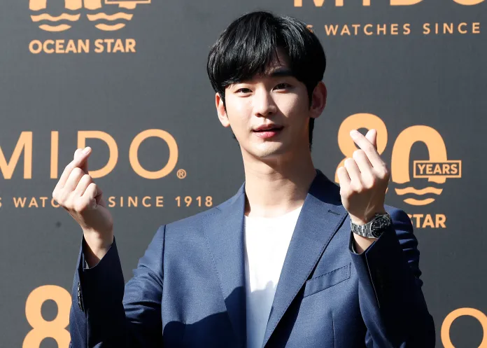 Kim Soo-hyun in a blue blazer and white shirt is posing for the camera, making a hand gesture with both hands, symbolizing love or affection, in front of a backdrop featuring the Mido logo for their 80th anniversary.