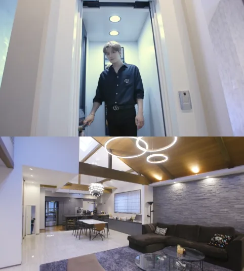 Kim Jaejoong standing at the entrance of an elevator, wearing a black shirt, with a modern living room visible in the background featuring high ceilings, a large window, and contemporary furniture.