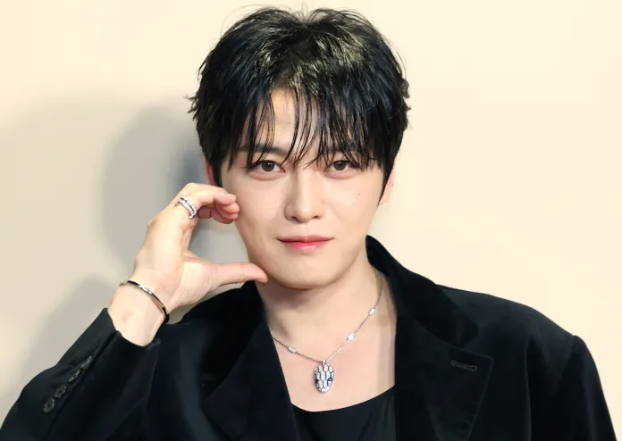Kim Jaejoong with dark, tousled hair and a stylish outfit poses with a hand on their chin, wearing silver jewelry against a neutral background.