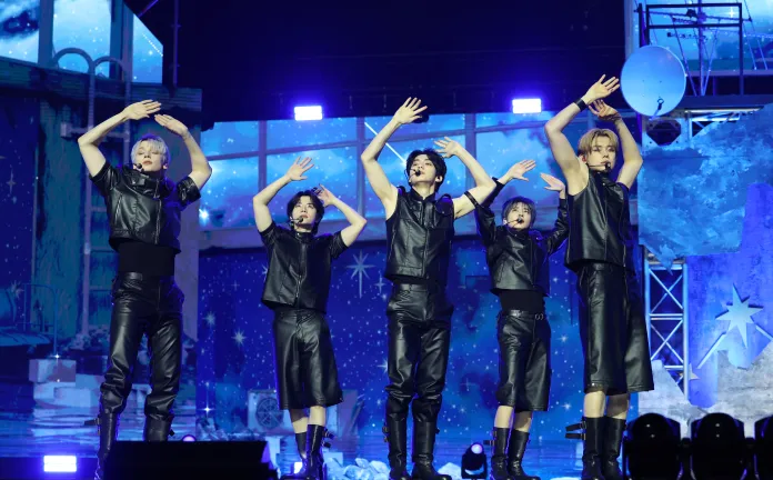 TOMORROW X TOGETHER performing on stage in matching black outfits, with arms raised, under colorful stage lighting.