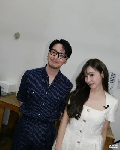 A man in a striped shirt and glasses stands next to a woman wearing a white dress, both smiling in a casual indoor setting.