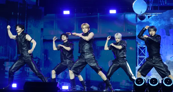TOMORROW X TOGETHER in stylish black outfits dancing energetically on stage with a vibrant light display in the background.