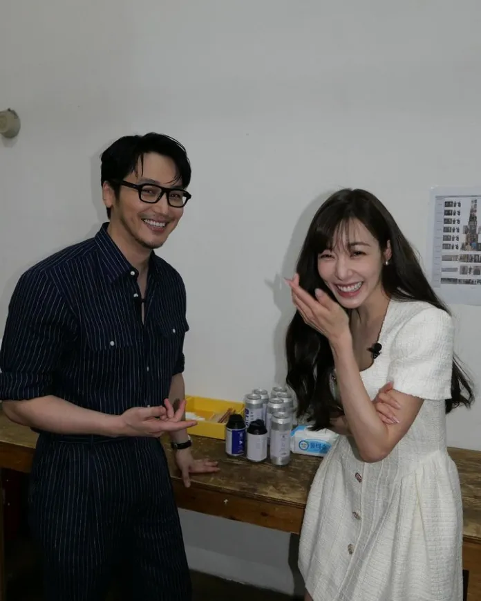 A man and a woman are smiling and interacting playfully in a casual indoor setting. The man is wearing glasses and a striped outfit, while the woman is dressed in a white, short-sleeved dress. They stand near a wooden table with various cans and items in the background.