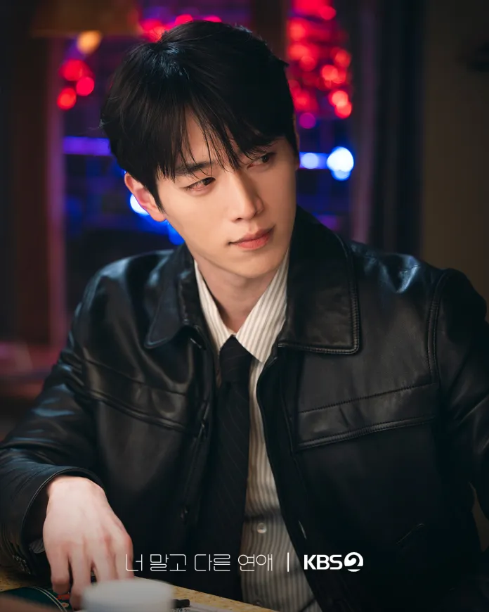 Seo Kang Joon wearing a black leather jacket and a white striped shirt, sitting at a table with a thoughtful expression, illuminated by colorful lights in the background.