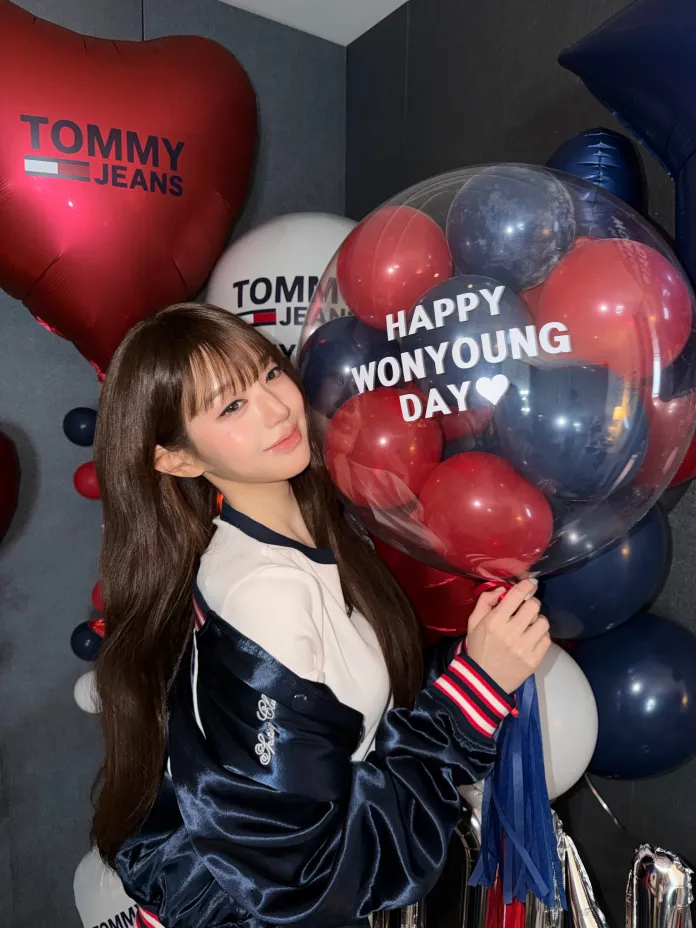 Wonyoung wearing a dark jacket poses with a large balloon that reads 'HAPPY WON YOUNG DAY' surrounded by colorful balloons and heart-shaped decorations in a festive setting.