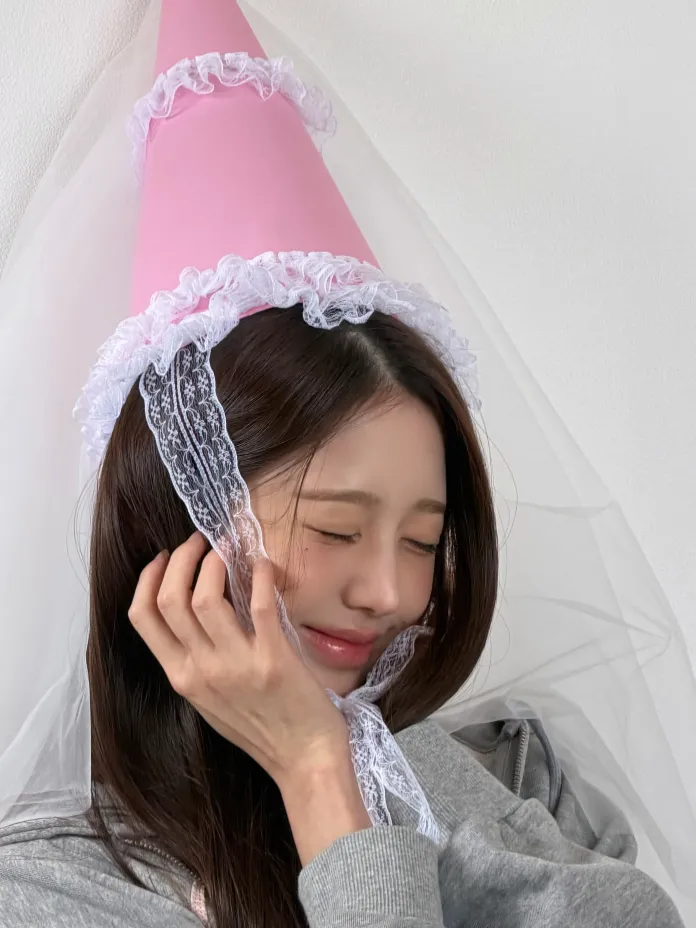 Wonyoung  wearing a pink party hat with lace trim and a veil, smiling and holding her face with one hand.