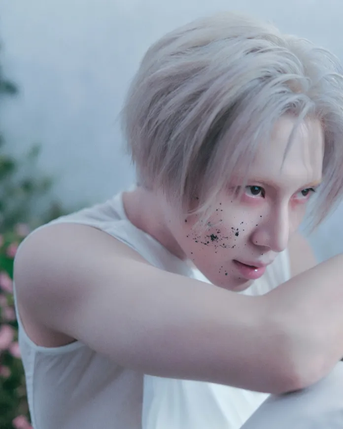 A close-up portrait ofT aemin with short, platinum blonde hair and a pale complexion, gazing intently with speckles of dirt on his face, positioned thoughtfully with arms crossed.