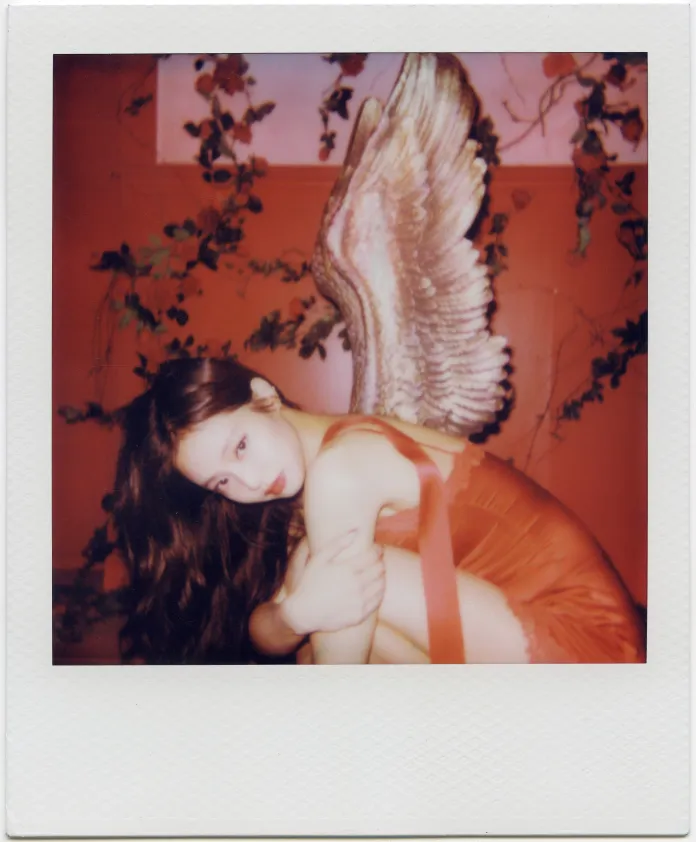 Jennie with long hair sits in a cozy pose, wearing an elegant dress. She has angelic wings behind her, set against a backdrop with trailing vines and soft colors.