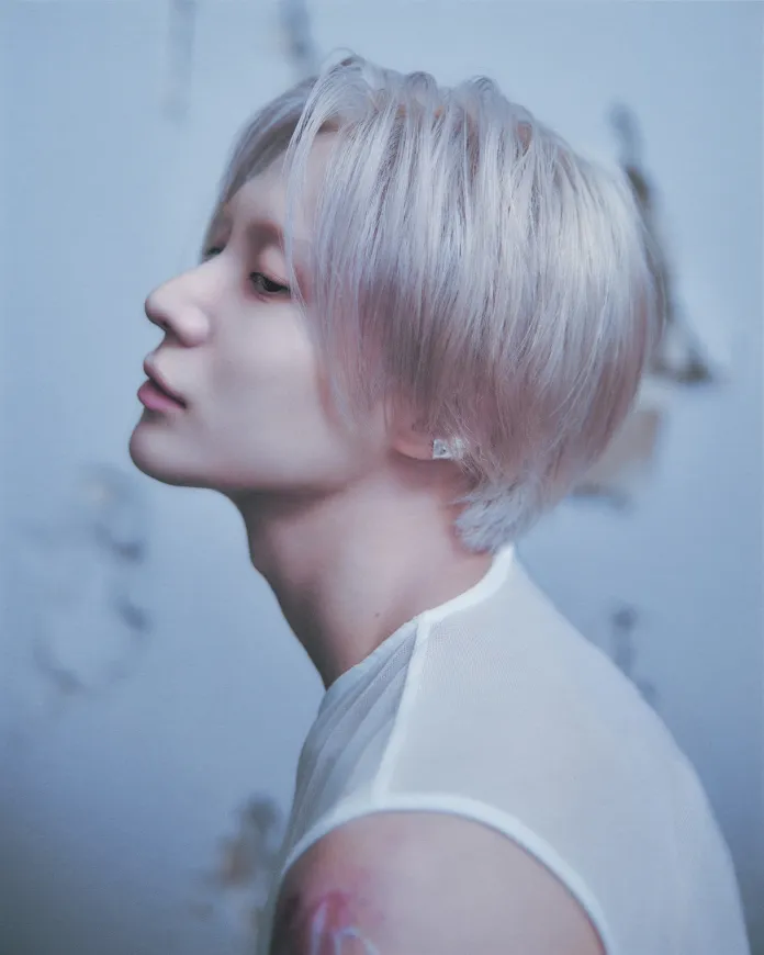 Profile view of Taemin with light blonde hair and a delicate facial structure, wearing a sheer top against a soft blue background.