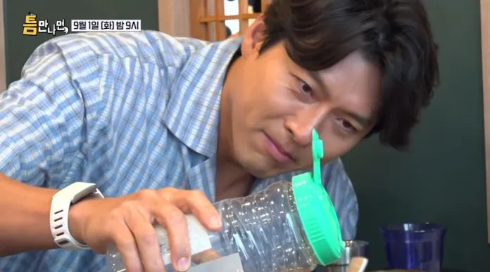 A man in a checkered shirt carefully pours liquid from a large container with a green lid while focusing intently on the task.