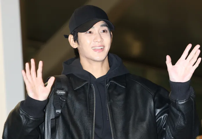 Kim Soo Hyun wearing a black baseball cap and a leather jacket smiles and waves at the camera, standing indoors.