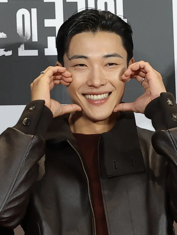 A smiling Woo Do Hwan showing a playful gesture with their hands near his face, wearing a brown leather jacket against a simple background.