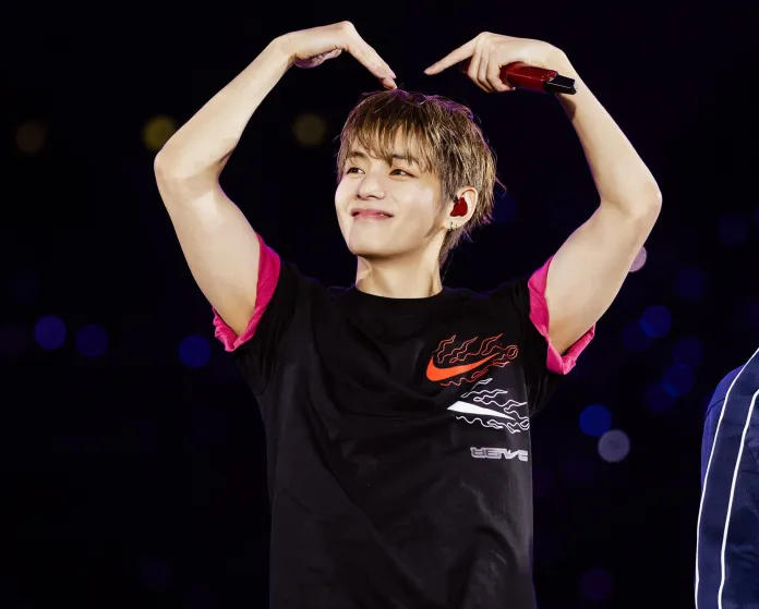 V wearing a black t-shirt with pink sleeves makes a heart shape with his hands, smiling at the audience. The background features blurred lights.