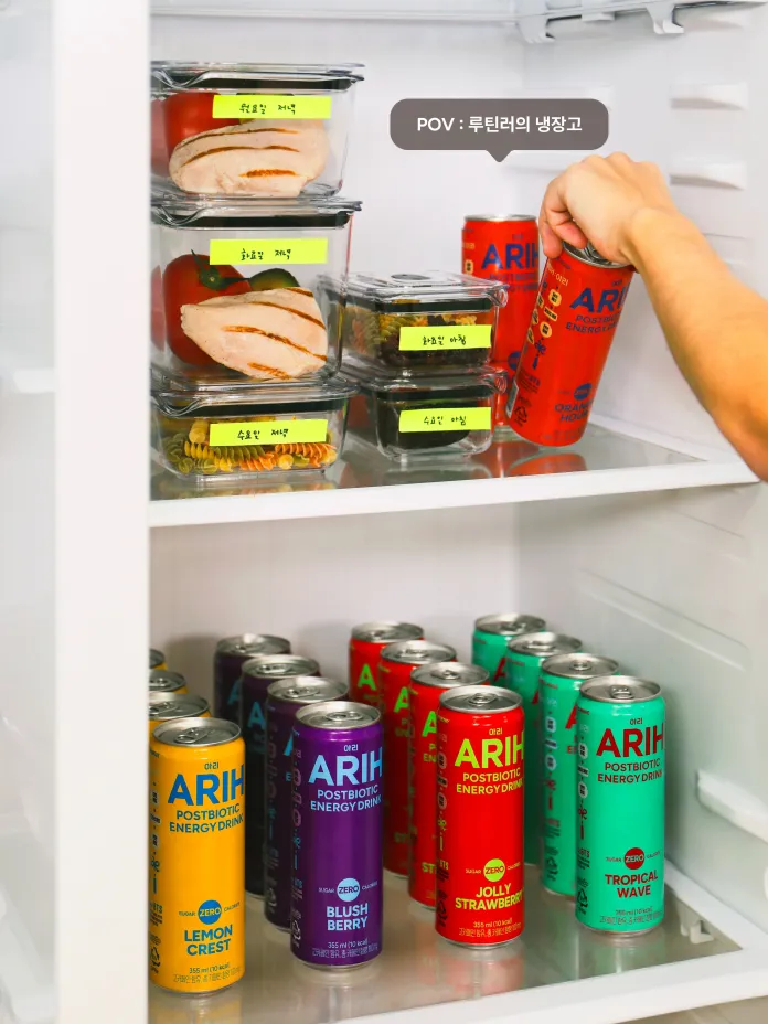 An open refrigerator filled with labeled food containers and colorful ARIH postbiotic energy drink cans. A hand is reaching for a red ARIH can.