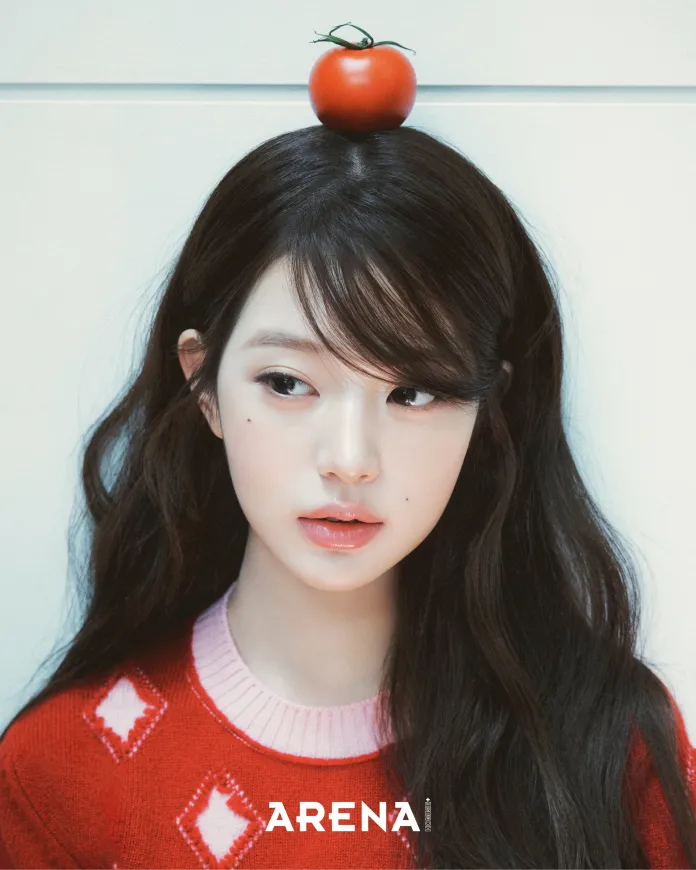 A close-up portrait of Jang Wonyoung with long dark hair, wearing a red sweater with a diamond pattern, and a tomato placed on top of her head.