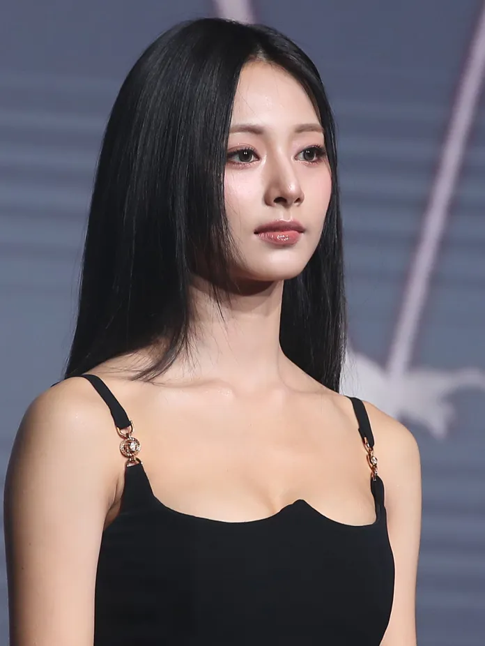 Close-up of Tzuyu with long black hair, wearing a black sleeveless top with decorative straps, looking serious.