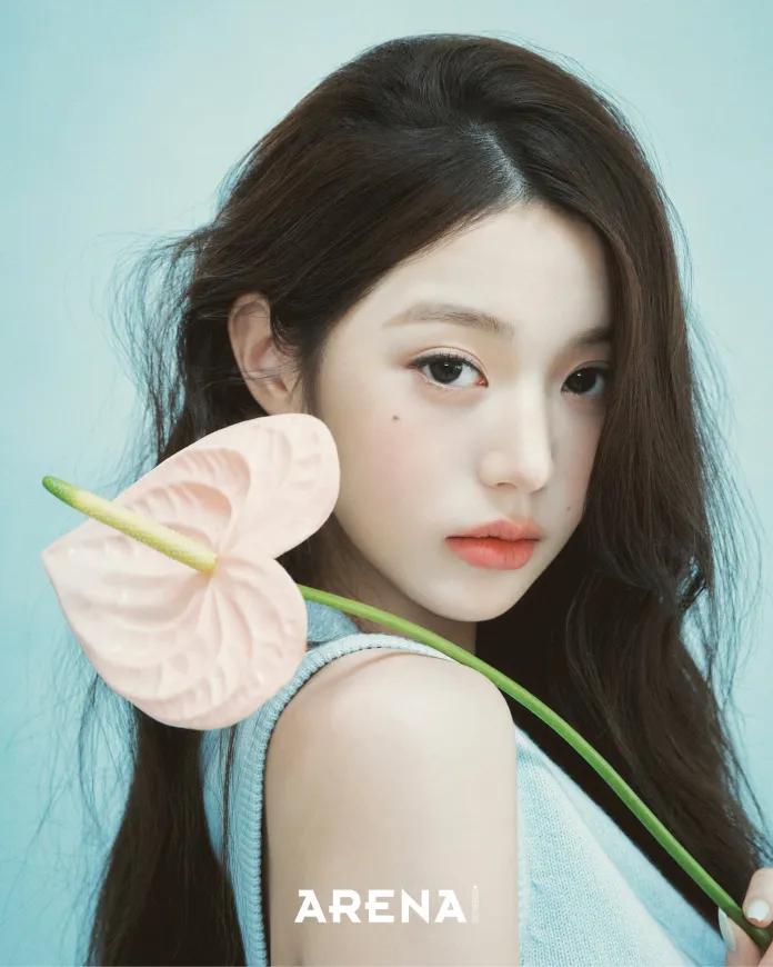 A close-up portrait of Jang Wonyoung with long dark hair, holding a large pink flower against a light blue background.