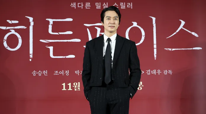 Song Seung Heon wearing a black suit with a white shirt stands confidently in front of a red backdrop of a promotional event. 