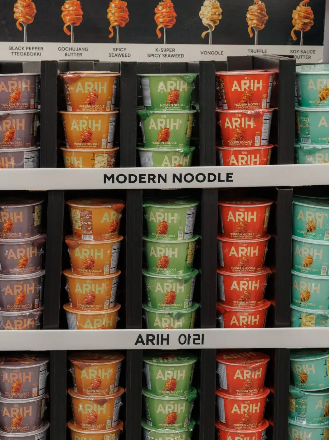 Display of various ARIH modern noodle cups in different colors and flavors on a shelf, with labels indicating product varieties.