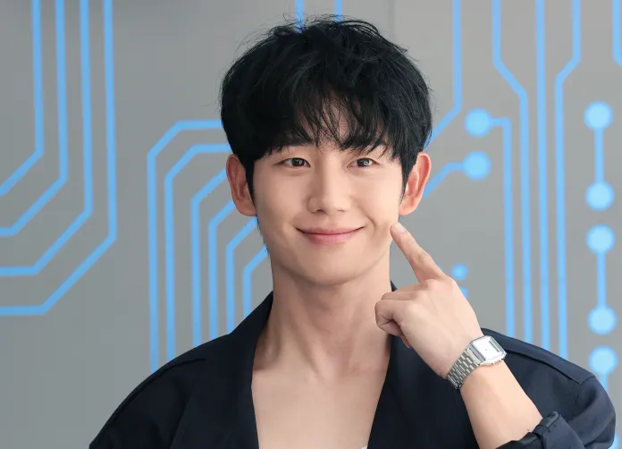 A smiling Jung Hae In with short black hair, wearing a dark jacket and a wristwatch, points at his cheek against a backdrop of blue circuit patterns.