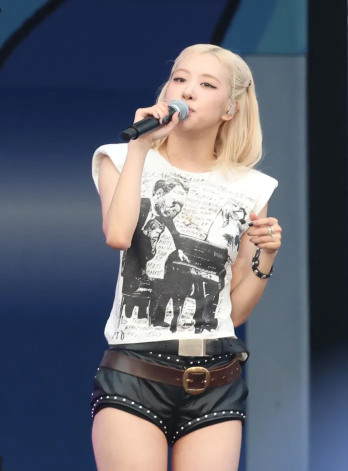 Rosé singing into a microphone while wearing a graphic t-shirt and leather shorts, with a stylish belt, on stage.