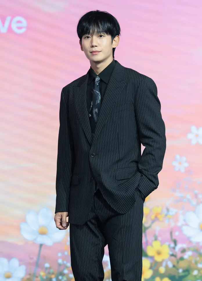 Jung Hae In in a striped black suit and tie stands confidently with his hands in his pockets, against a colorful floral backdrop.