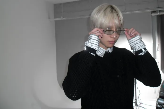 TAEMIN with short blonde hair wearing glasses, a black sweater, and a striped shirt, adjusting their glasses in an indoor setting.