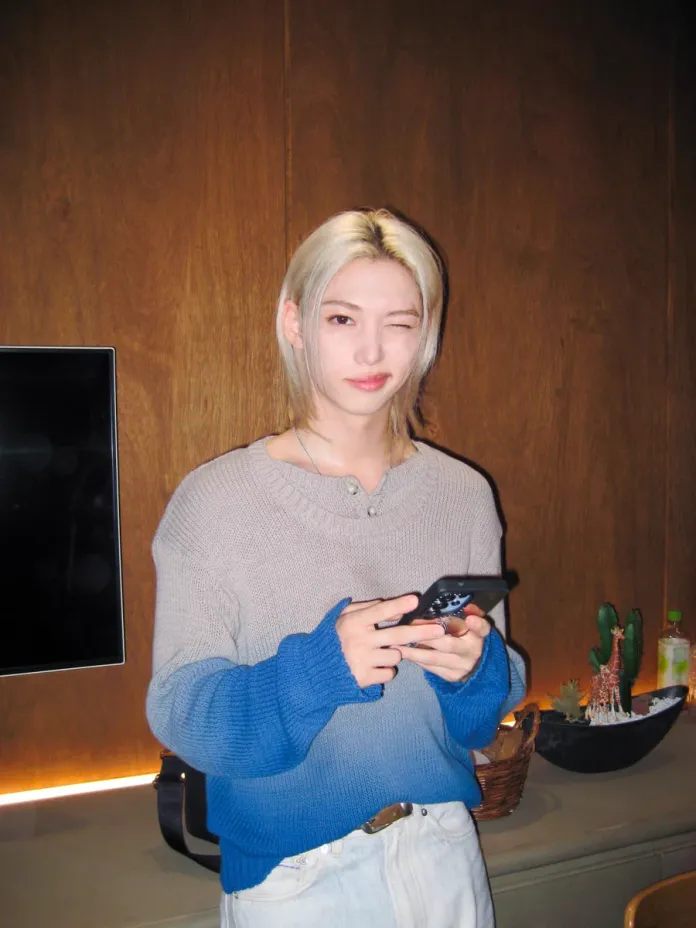 A person with blonde hair wearing a gray and blue sweater winks while holding a smartphone, standing in front of a wooden wall with decorative plants in the background.