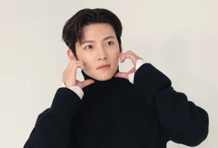 Ji Chang Wook in a black turtleneck sweater, holding his face with both hands and making a heart shape with his fingers, looking thoughtfully at the camera.